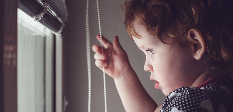 Child Looking Out Window Holding Blind Cord 